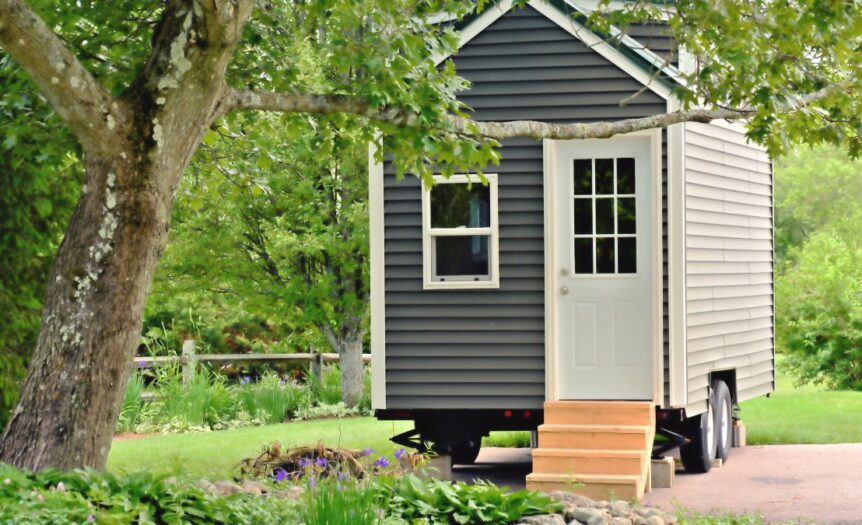 The exterior of a tiny home on wheels with gray siding, white trim, wooden front steps, and a white front door.