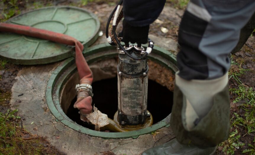 A close-up of a worker with rubber boots and gloves lowering equipment through a sewer hatch for a sewer line repair.