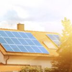 Several solar panels sit on an orange-tiled roof, creating a rectangle. The sun shines through a tree next to the home.