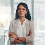 A confident businesswoman stands in a corporate office, arms crossed and smiling in front of a conference room.