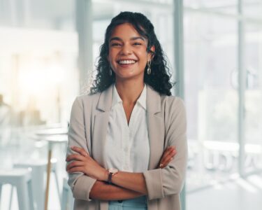 A confident businesswoman stands in a corporate office, arms crossed and smiling in front of a conference room.