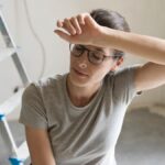 A woman in a gray T-shirt wipes her forehead while kneeling in a room with unfinished floors and walls.