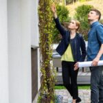 A real estate agent is examining the exterior of the home alongside an inspector, who is holding blueprints in his hand.