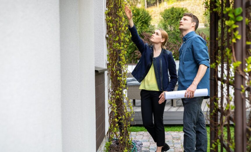 A real estate agent is examining the exterior of the home alongside an inspector, who is holding blueprints in his hand.
