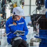 A woman kneels near a black and blue factory machine, inspecting it. She holds a tablet and wears protective gear.