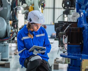 A woman kneels near a black and blue factory machine, inspecting it. She holds a tablet and wears protective gear.