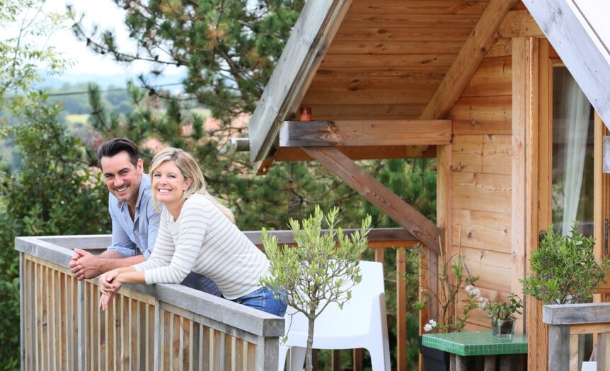 A smiling man and a woman stand on the wooden deck of a cabin, leaning on the railing. Trees appear in the background.