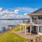 A wide-angle view of a grey, two-story, modern lakefront home, with two docks on the water's edge.