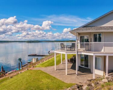 A wide-angle view of a grey, two-story, modern lakefront home, with two docks on the water's edge.