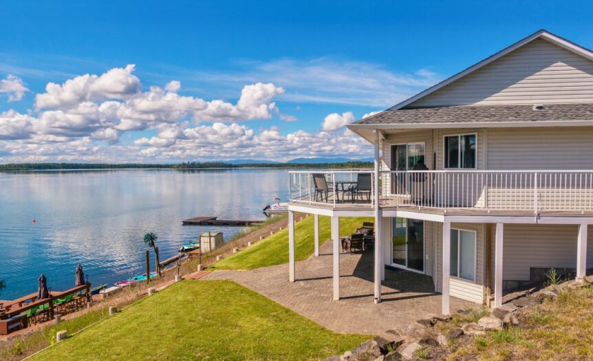 A wide-angle view of a grey, two-story, modern lakefront home, with two docks on the water's edge.