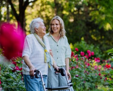 An adult granddaughter is assisting her grandmother as they walk in the park, with the grandmother using a walker.