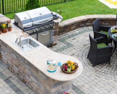 A backyard kitchen with a concrete brick island, sink, and grill installed next to a round table with chairs.