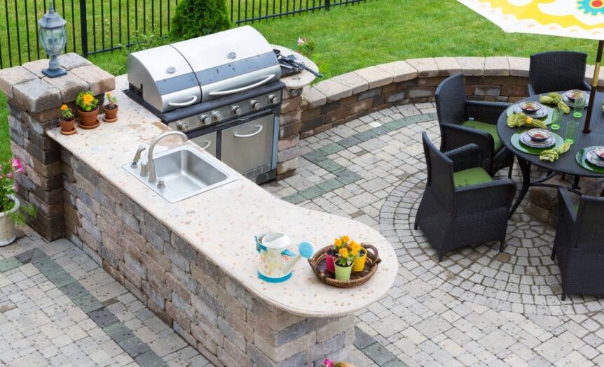 A backyard kitchen with a concrete brick island, sink, and grill installed next to a round table with chairs.