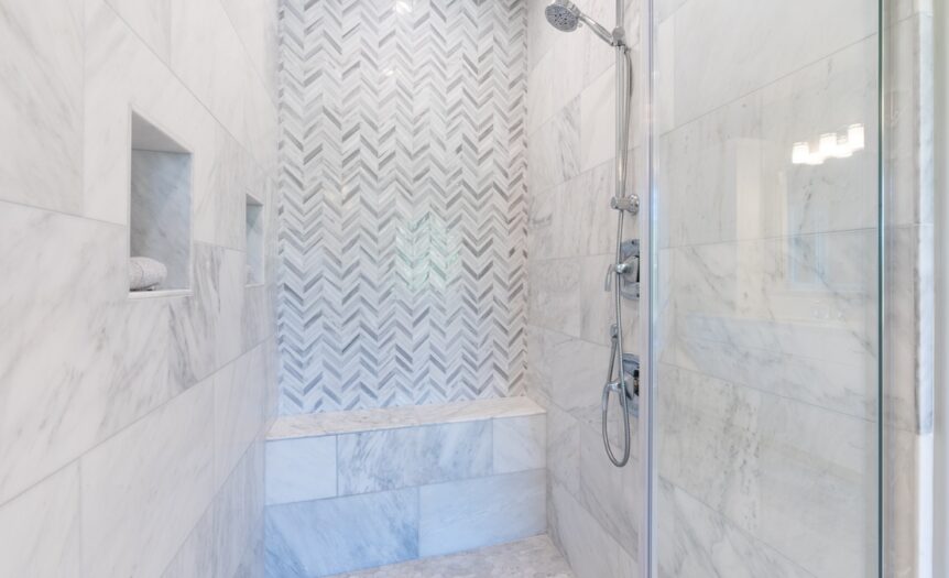 A walk-in shower with white and light gray tiles and an open glass door, a built-in tile bench, and a handheld showerhead.