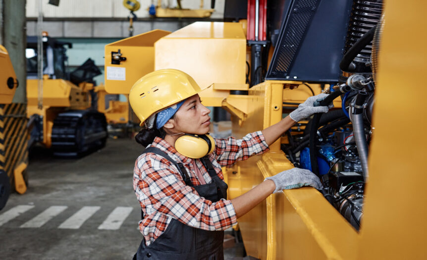 A young woman wearing a plaid shirt and industrial PPE inspects the components of a piece of heavy equipment painted yellow.