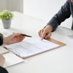 Two people sit across from each other at a white desk, both gesturing to a clipboard with documents on it.