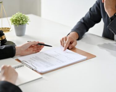 Two people sit across from each other at a white desk, both gesturing to a clipboard with documents on it.