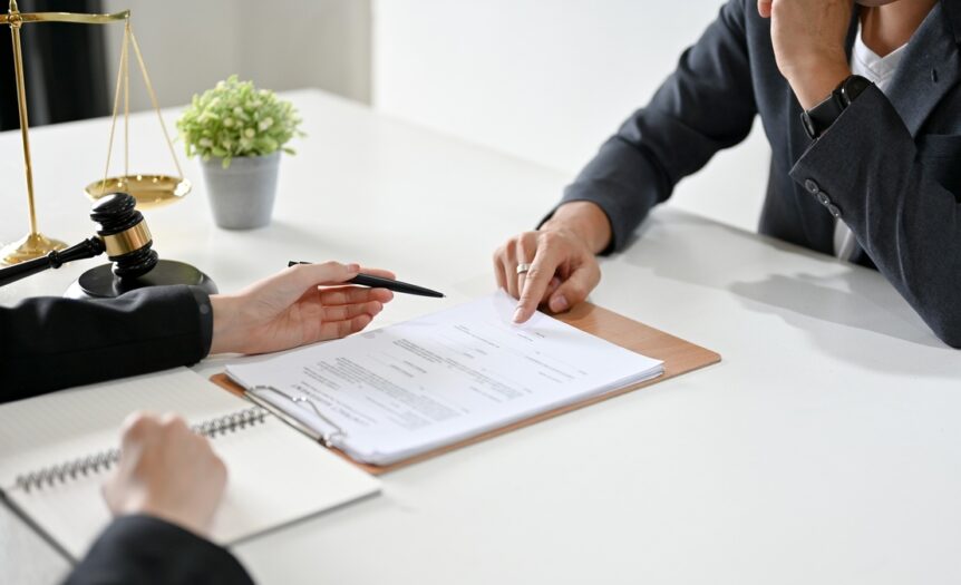 Two people sit across from each other at a white desk, both gesturing to a clipboard with documents on it.