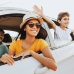 A family of three smiling in a car. The child is in the backseat, leaning out the window with their hands outstretched.