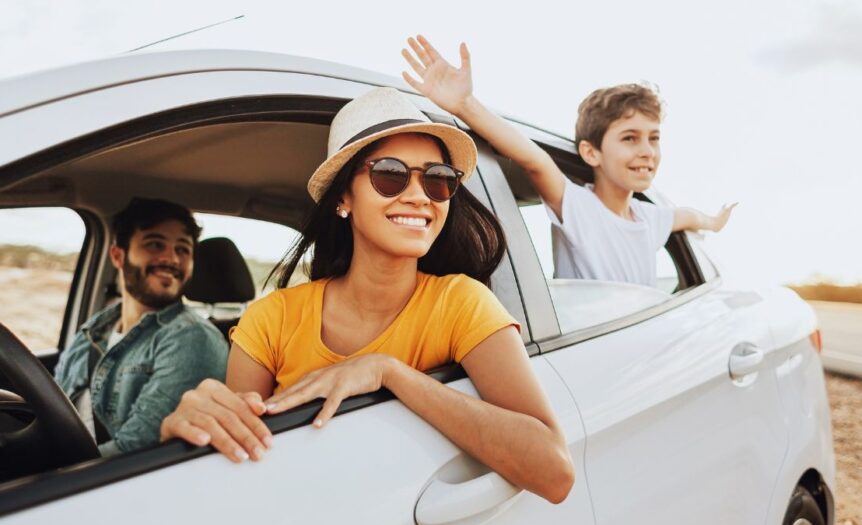 A family of three smiling in a car. The child is in the backseat, leaning out the window with their hands outstretched.