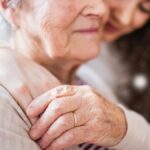 A seated, gray-haired woman with closed eyes peacefully receiving a warm one-armed hug from a younger woman.