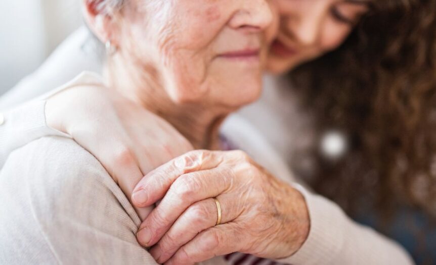 A seated, gray-haired woman with closed eyes peacefully receiving a warm one-armed hug from a younger woman.
