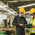 Two men in gas masks and hard hats look at clipboards while standing inside a warehouse strewn with wreckage.