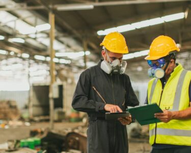 Two men in gas masks and hard hats look at clipboards while standing inside a warehouse strewn with wreckage.