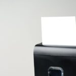 A small, black paper shredder is shredding a plain, white piece of paper. The background is cream-colored.