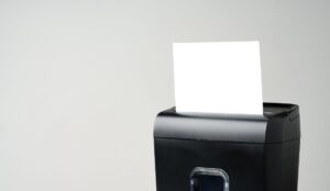 A small, black paper shredder is shredding a plain, white piece of paper. The background is cream-colored.