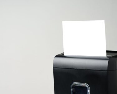 A small, black paper shredder is shredding a plain, white piece of paper. The background is cream-colored.