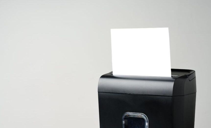 A small, black paper shredder is shredding a plain, white piece of paper. The background is cream-colored.
