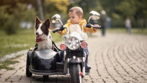 A kid sits in a toy replica of a motorcycle in a park. His dog sits in the vehicle's sidecar and wears a bandana.