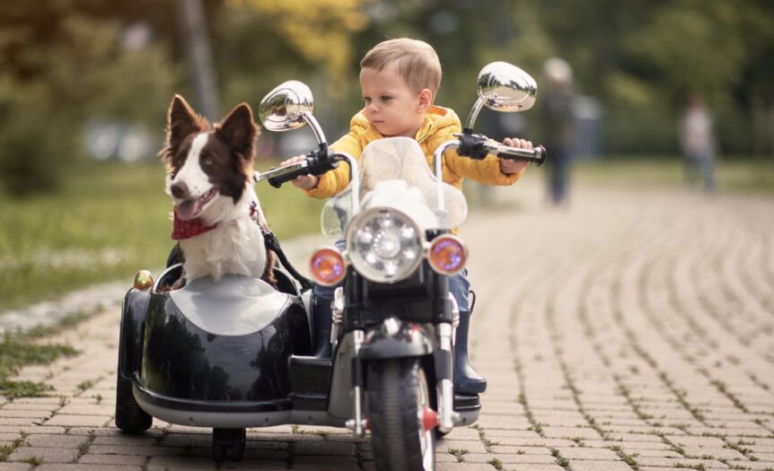 A kid sits in a toy replica of a motorcycle in a park. His dog sits in the vehicle's sidecar and wears a bandana.