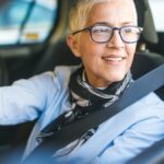 A smiling, middle-aged woman with short hair and glasses sitting in a black car with her hands on the wheel.