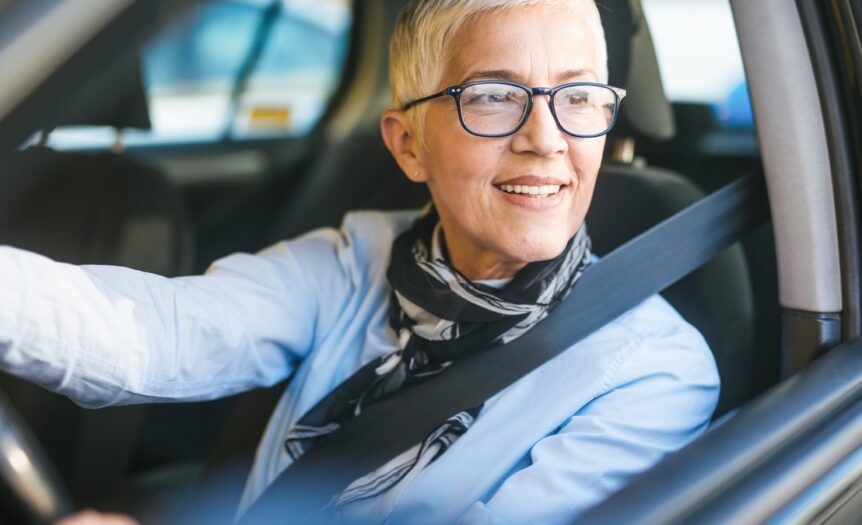 A smiling, middle-aged woman with short hair and glasses sitting in a black car with her hands on the wheel.