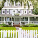 A large house with a wrap-around porch on the first floor. There are trees surrounding it and a white front gate.