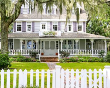 A large house with a wrap-around porch on the first floor. There are trees surrounding it and a white front gate.