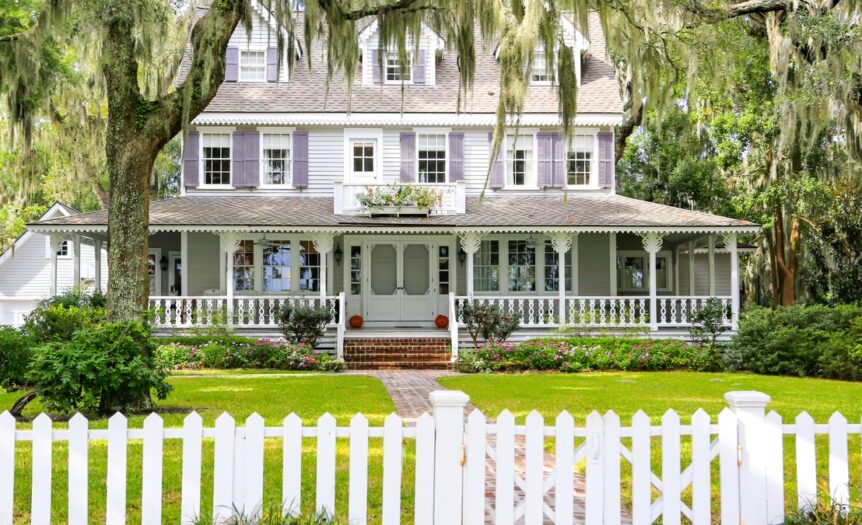 A large house with a wrap-around porch on the first floor. There are trees surrounding it and a white front gate.