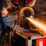 A worker wearing protective gloves uses a grinder to cut through a large piece of metal in a workshop.
