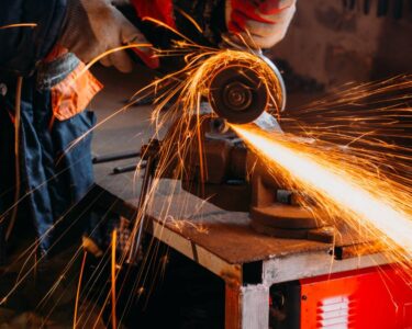 A worker wearing protective gloves uses a grinder to cut through a large piece of metal in a workshop.