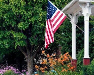 An American flag hangs off the end of a long white flagpole attached to the pillar on the front porch of a house.