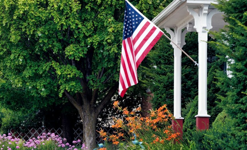 An American flag hangs off the end of a long white flagpole attached to the pillar on the front porch of a house.