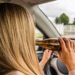 A woman sits in the driver's seat of a car, with one hand on the steering wheel and another holding a beer bottle.