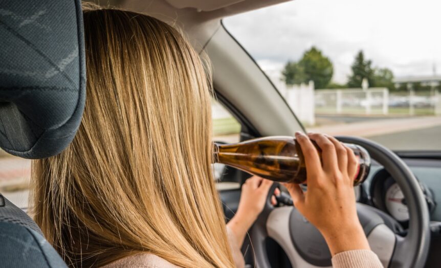A woman sits in the driver's seat of a car, with one hand on the steering wheel and another holding a beer bottle.