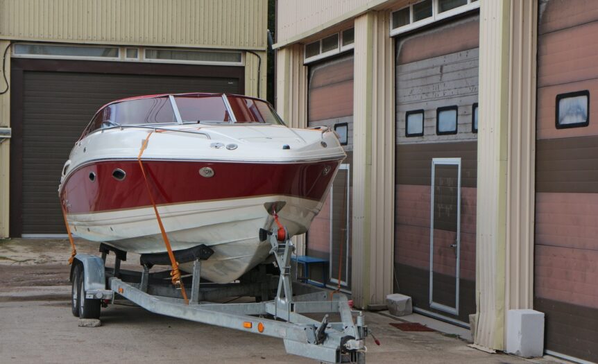 A red and white boat secured to a silver trailer with orange straps sits in front of a brown garage door.