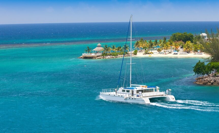 An aerial view of a white catamaran sailing past a beach with palm trees and a gazebo on a sunny day.