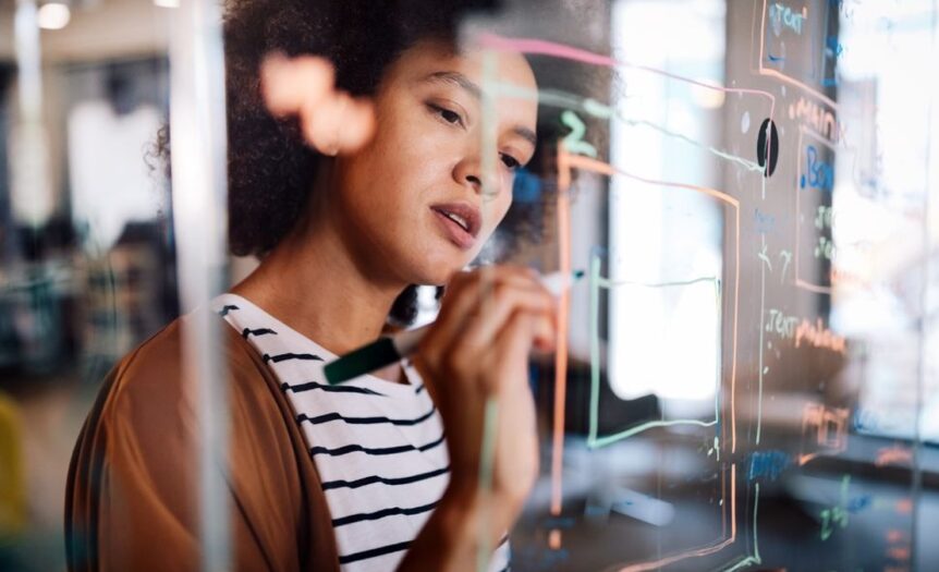 A Black woman is concentrating and writing on the glass board with colorful chalk pens in her office.