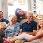 Two young children smile as they sit on a couch with three middle aged adults and two elderly people.