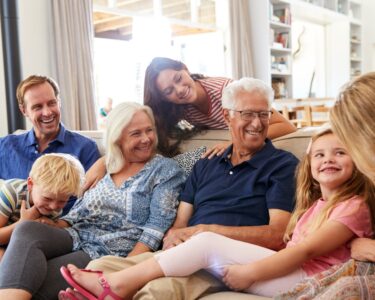 Two young children smile as they sit on a couch with three middle aged adults and two elderly people.